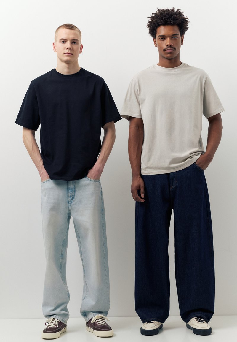 Deux hommes se tiennent côte à côte sur un fond blanc, vêtus de t-shirts amples unis et de jeans à jambes larges, les mains dans les poches.