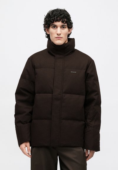 Brūna pūpera jaka ar augstu apkakli, izšūtiem paneļiem un logo izšūtu krūtīs. Izgatavota no mīksta, teksturēta materiāla.