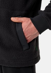 Giacca in fleece nera con superficie texture, polsini regolabili e tasca laterale. Presenta un dettaglio logo sottile e un bordo a contrasto.