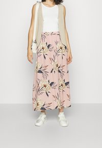 Gonna maxi rosa con motivo floreale, tessuto morbido, vita elasticizzata, abbinata a una maglietta bianca senza maniche e un gilet in maglia, completata da sneakers bianche.