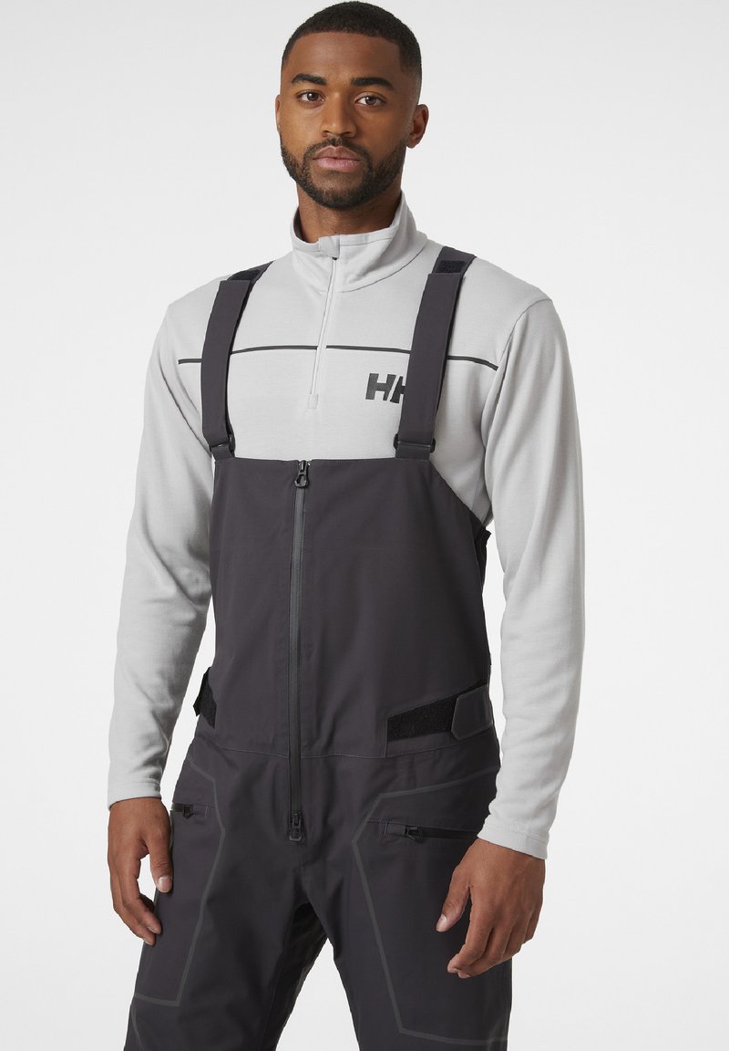 Overalls de esquí impermeables de color gris oscuro con tirantes ajustables, cierre frontal de cremallera y bolsillos con cremallera. Llevados sobre una camiseta de manga larga gris claro.