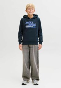 Sudadera con capucha azul marino con bolsillo delantero, con texto en blanco y azul; combinada con vaqueros de pierna ancha gris claro y zapatillas grises.