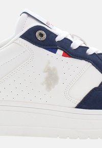 Zapatillas blancas con acentos de gamuza en color marino, cuero perforado y un logo sutil. Incluyen un ojal de metal y un detalle de bandera tricolor.