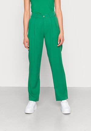 Mujer con pantalones plisados de talle alto en color verde brillante, con cierre de botón y cremallera frontal, combinados con zapatillas blancas de plataforma.