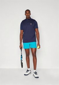 Tmavě modré polo tričko se zipem u límce, v kombinaci s jasně tyrkysovými šortkami. Drží tenisovou raketu a míček. Bílé sportovní boty s tmavými detaily.