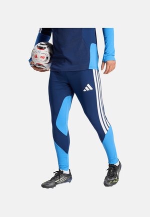 Persona con abbigliamento sportivo blu e tacchetti neri tiene una palla da calcio Adidas bianca e rossa su uno sfondo semplice.