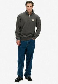 Grijze ritspullover met een hoge kraag en geborduurd logo, gecombineerd met losse blauwe denim jeans en zwarte leren schoenen.