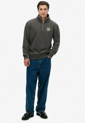 Pullover grigio con zip e collo alto, arricchito da un logo ricamato, abbinato a jeans blu larghi e scarpe in pelle nere.