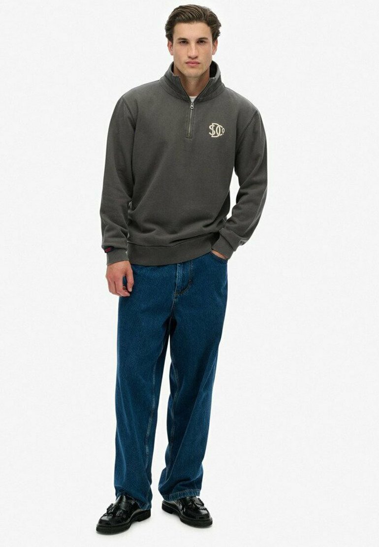 Grijze ritspullover met een hoge kraag en geborduurd logo, gecombineerd met losse blauwe denim jeans en zwarte leren schoenen.