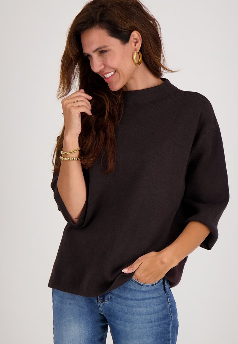 Donkerbruine oversized trui met een hoge hals en 3/4 mouwen, gedragen met een blauwe spijkerbroek. Gladde textuur, minimalistisch ontwerp.