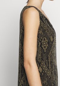 Kvinna i en ärmlös svart och olivgrön klänning med ett geometriskt mönster, sedd från sidan mot en enkel ljus bakgrund.