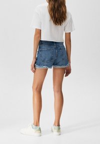 Shorts de mezclilla con bajos deshilachados, de color azul oscuro, con dos bolsillos traseros, combinados con zapatillas blancas. Lleva una camiseta corta blanca.