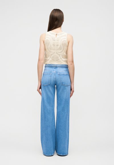 Γυναίκα με μακριά καστανά μαλλιά που φοράει αμάνικη μπεζ πλεκτή μπλούζα και μπλε τζιν παντελόνι φαρδύ με ψηλή μέση, στέκεται γυρισμένη προς τα πίσω.