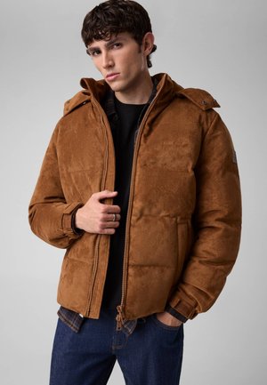 Ung mand iført en brun polstret jakke med hætte, sort skjorte og blå jeans, poserer med den ene hånd nær lynlåsen.
