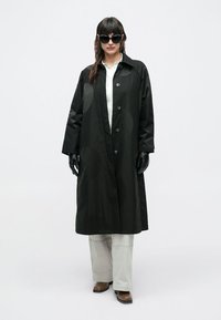 Palton lung negru cu textură netedă, prevăzut cu închidere cu nasturi și guler larg. Purtat peste o cămașă deschisă la culoare și pantaloni deschiși.