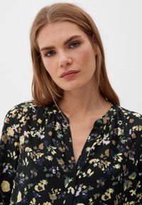 Schwarze Bluse mit floralem Muster aus leichtem, transparentem Stoff. Merkmale sind bunte Blumendrucke in Gelb, Lila und Grün. Knopfverschluss vorne und Faltenlegung.
