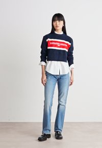 Navy sweater with red and white stripes, featuring "tommy jeans" logo; layered over a white collared shirt. Light blue jeans and black shoes.