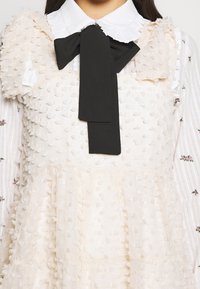 Robe blanche texturée avec des pois en relief sur un chemisier blanc à petits imprimés floraux et un grand nœud papillon noir au col.