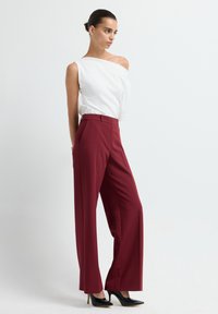 Blouse blanche à épaules dénudées associée à un pantalon bourgogne taille haute et à larges jambes, et des escarpins noirs à bout pointu, sur un fond neutre.