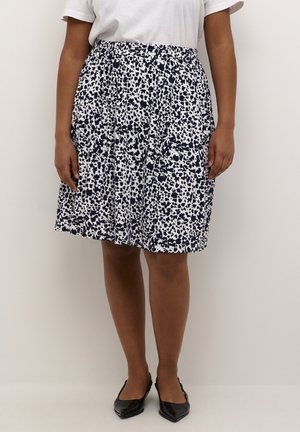 Kvinde iført et knælængde hvidt og sort plettet nederdel, hvid top og sorte flade sko med spids tå, stående mod en ensfarvet baggrund.