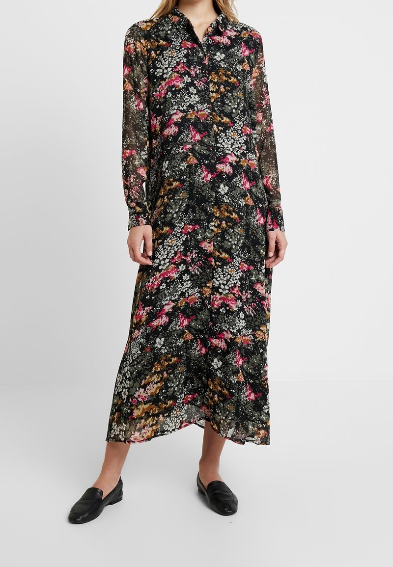 Femme portant une robe noire à manches longues boutonnée avec un imprimé floral rose, blanc et marron, associée à des chaussures noires sans lacets.