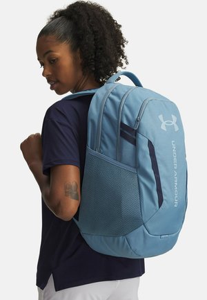 Light blue backpack with mesh side pocket, padded straps, and Under Armour logo in gray. Features a streamlined design with multiple compartments.