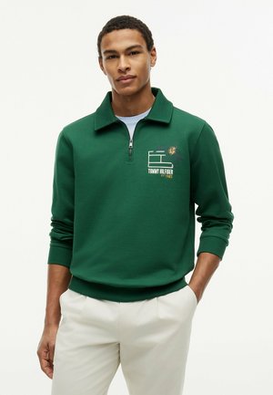 Mężczyzna ubrany w zieloną bluzę z zamkiem ćwiartkowym i logo Tommy Hilfiger oraz kremowe spodnie, stoi z jedną ręką w kieszeni na białym tle.