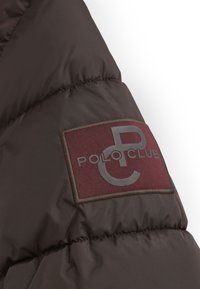 Sección de chaqueta acolchada marrón con una superficie texturizada. Presenta un parche de logo en burdeos con "POLO CLUB" y una "P" estilizada en gris.