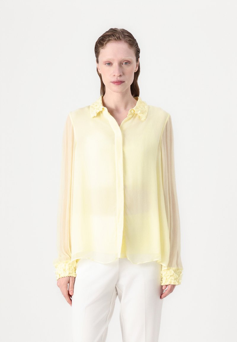Chiffon-Bluse in sanftem Gelb mit floralen Akzenten am Kragen und gerafften Manschetten, aus leichtem, halbtransparentem Stoff und mit Knopfverschluss.