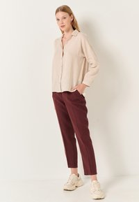 Camicia button-up in lino di un leggero beige abbinata a pantaloni tapered borgogna, indossata con sneakers beige. Design semplice e vestibilità rilassata.