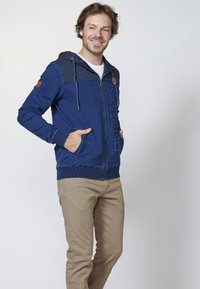 Blue zip-up hoodie with a textured upper section, drawstring hood, and two front pockets. Features ribbed cuffs and a logo on the front.