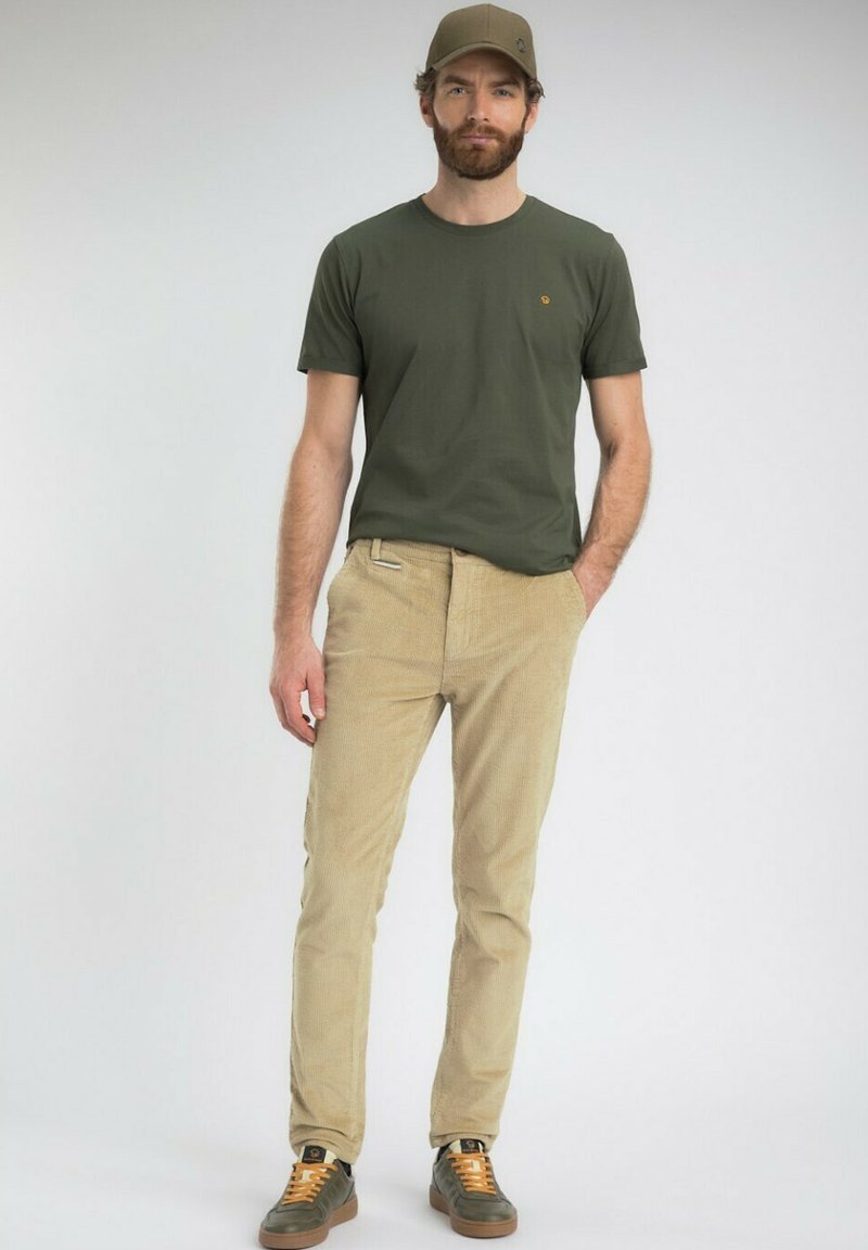 Homme portant un t-shirt vert olive, un pantalon en velours côtelé beige, des baskets vert olive avec des lacets orange, et une casquette assortie verte olive, se tenant avec les mains dans les poches.