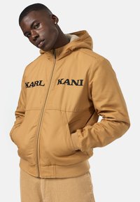 Brown hooded jacket featuring embroidered "KARL" and "KANI" logos, zip closure, and ribbed cuffs and hem. Soft, textured inner lining.