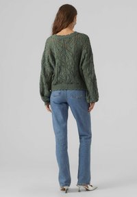 Grüner, gestrickter Pullover mit komplexen Spitzenmustern, lockerer Passform, geripptem Saum, kombiniert mit hellblauen Jeans und weißen Absätzen.