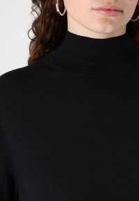 Maglione a collo alto di colore nero realizzato in un morbido tessuto a coste, con un collo alto e una silhouette aderente. Inquadratura ravvicinata del collo e della parte superiore del corpo.