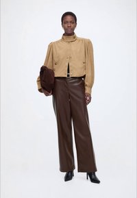 Γυναίκα που φοράει μπεζ μπλούζα με ψηλό λαιμό και φουσκωτά μανίκια, καφέ παντελόνι από δέρμα με φαρδιά πόδια, μαύρα μποτάκια αστραγάλου, κρατώντας καφέ τσάντα.