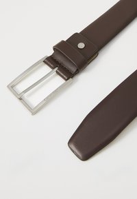 Cinturón de cuero marrón con hebilla rectangular plateada. Textura suave con un extremo puntiagudo. Presenta un logotipo de la marca en la hebilla.
