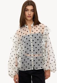 Femme portant un chemisier blanc transparent à pois noirs, boutonné devant, manches à volants, et un pantalon noir devant un fond uni.