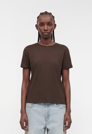Kvinde med flettet hår iført en ensfarvet mørkebrun kortærmet skjorte og lyseblå jeans, der står mod en hvid baggrund.
