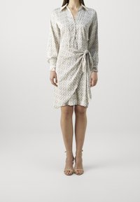 Femme portant une robe longue à manches longues à motifs beige avec col et détail de nœud, associée à des escarpins à bride de cheville beige.