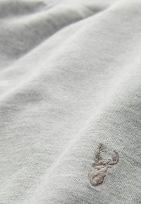 Hellgrauer Stoff mit einer subtilen Textur, mit einem gestickten dunkelgrauen Hirschmotiv in der unteren linken Ecke.
