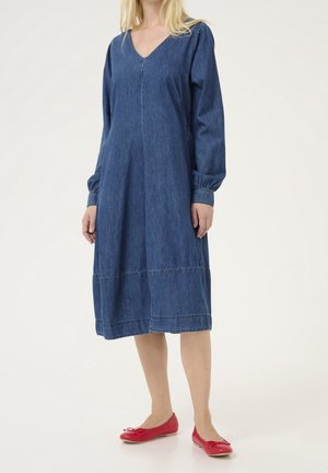 Vrouw draagt een denimjurk met lange mouwen tot op de knie, een V-hals en rode ballerina's, staand tegen een effen achtergrond.