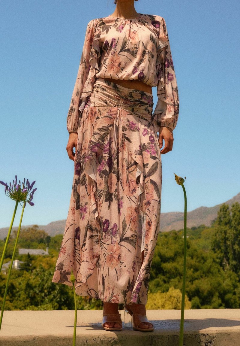 Blomstermønstret sæt bestående af en langærmet cropped top og en maxiskørt i blød pink, med grønne planter i baggrunden.