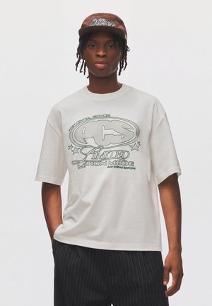 Junger Mann mit geflochtenem Haar, der eine braune Mütze und ein weißes übergroßes T-Shirt mit grafischem Text sowie schwarze Nadelstreifenhosen trägt.