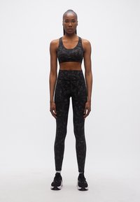 Γυναίκα που στέκεται φορώντας μαύρο αθλητικό σουτιέν με μάρμαρο σχέδιο, αντίστοιχο υψηλόμεσο κολάν, λευκές κάλτσες και μαύρα αθλητικά παπούτσια σε λευκό φόντο.