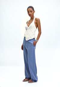 Lichtbeige haltertop met verticale strepen, gecombineerd met losse lichtblauwe broek. Model draagt tan sandalen en een zilveren armband.