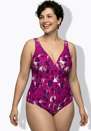 Smiling woman with short curly hair wearing a pink and purple patterned one-piece swimsuit with a V-neckline.