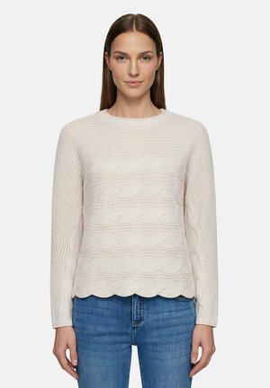 Femme aux cheveux bruns lisses portant un pull torsadé beige clair et un jean bleu, debout devant un fond uni.