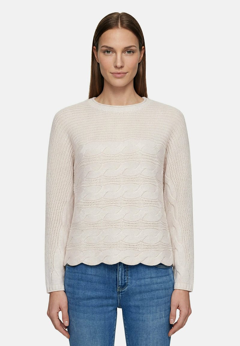 Femme aux cheveux bruns lisses portant un pull torsadé beige clair et un jean bleu, debout devant un fond uni.