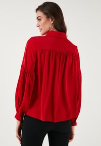 Red blouse with a collared neck, puffed long sleeves, and gathered details at the back. Made of smooth fabric, it has a loose fit.